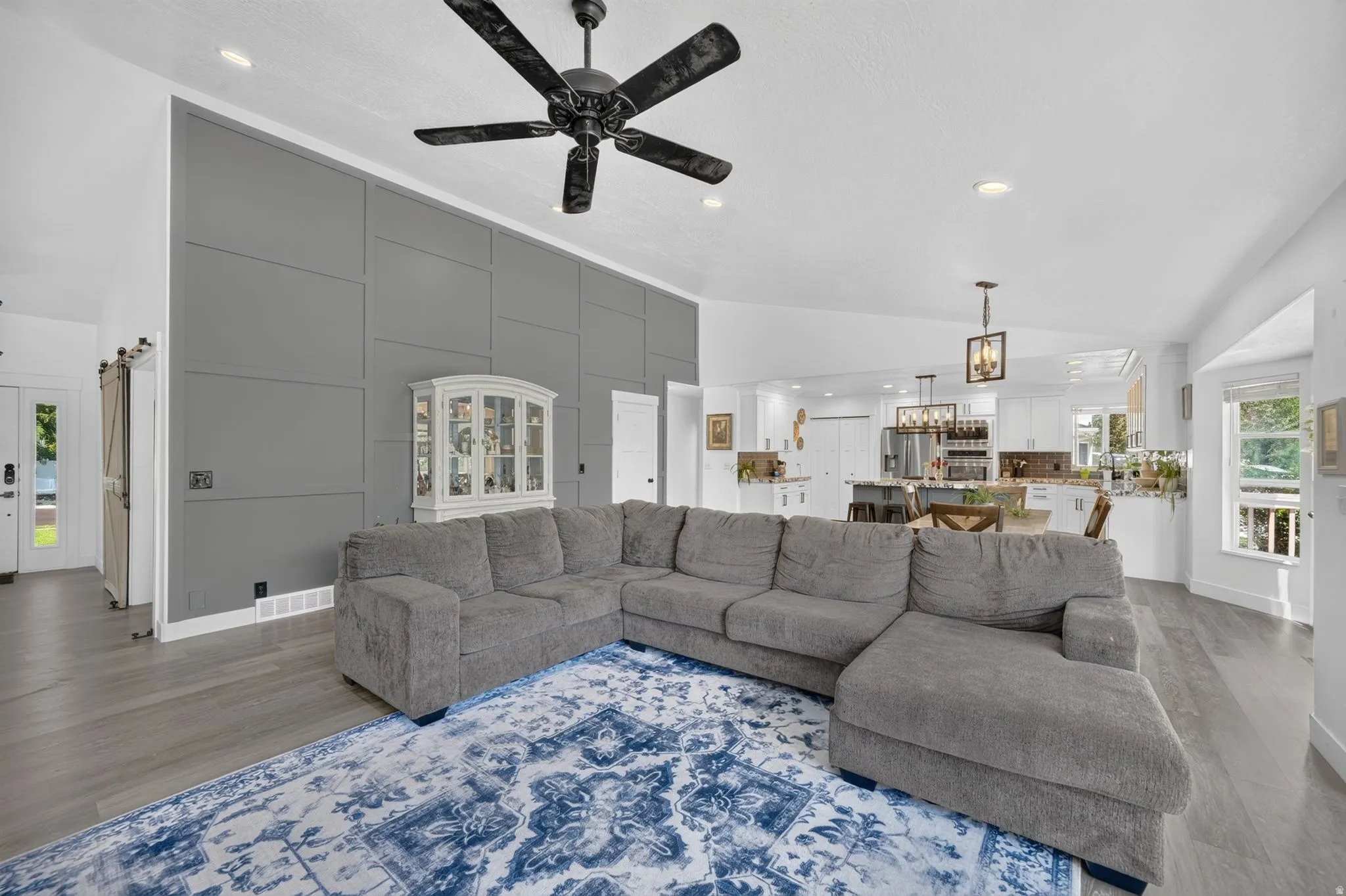 Vaulted ceilings with recessed lighting, wood-finish flooring, accent wall with decorative paneling, black ceiling fan, and a kitchen with white cabinetry