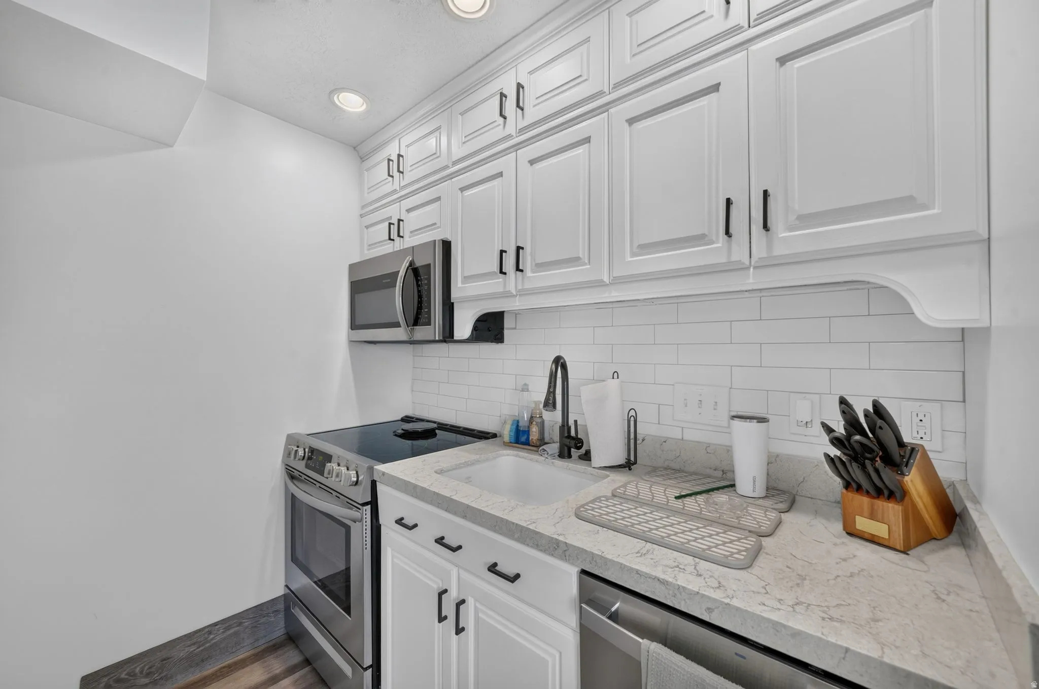 White shaker-style cabinetry with matte black hardware, stainless steel appliances, subway tile backsplash, light-toned countertops, and wood-finish flooring