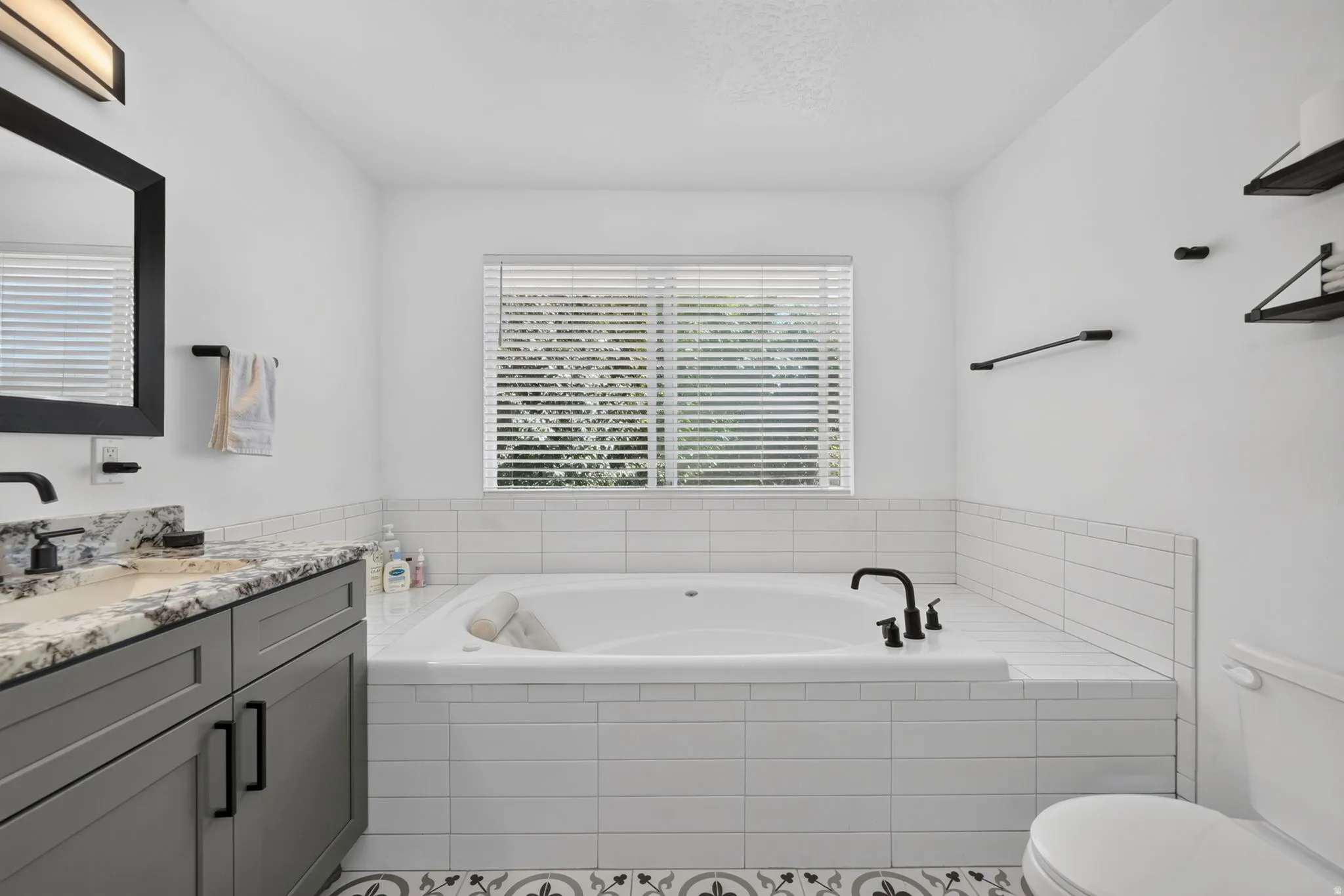 Bathroom featuring a large built-in soaking tub, a single vanity with a stone countertop, a window with blinds, and decorative floor tiling