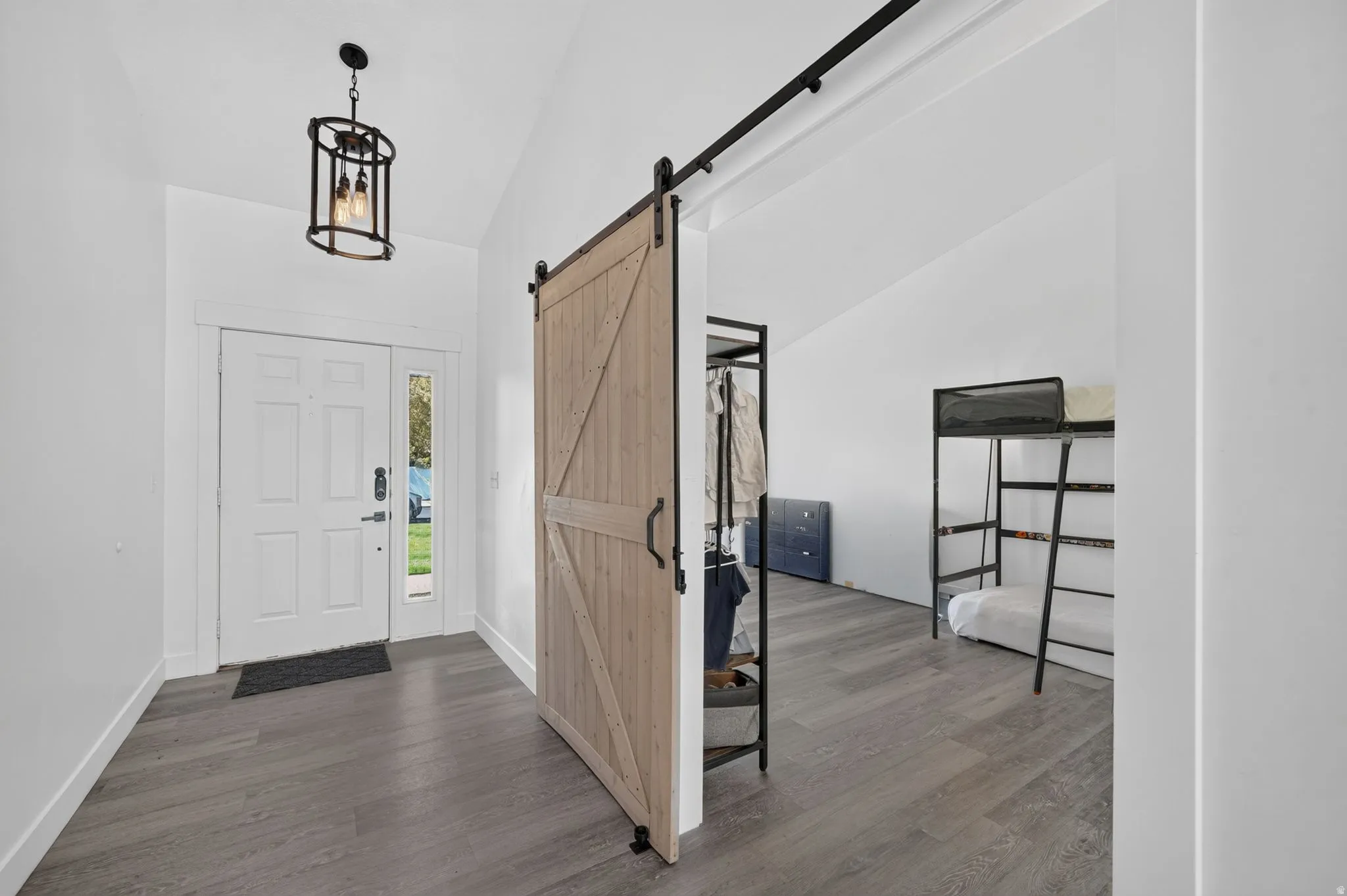 Vaulted ceiling, wood-finish flooring, white door with sidelight, black metal pendant light fixture, and a light wood-finish barn door