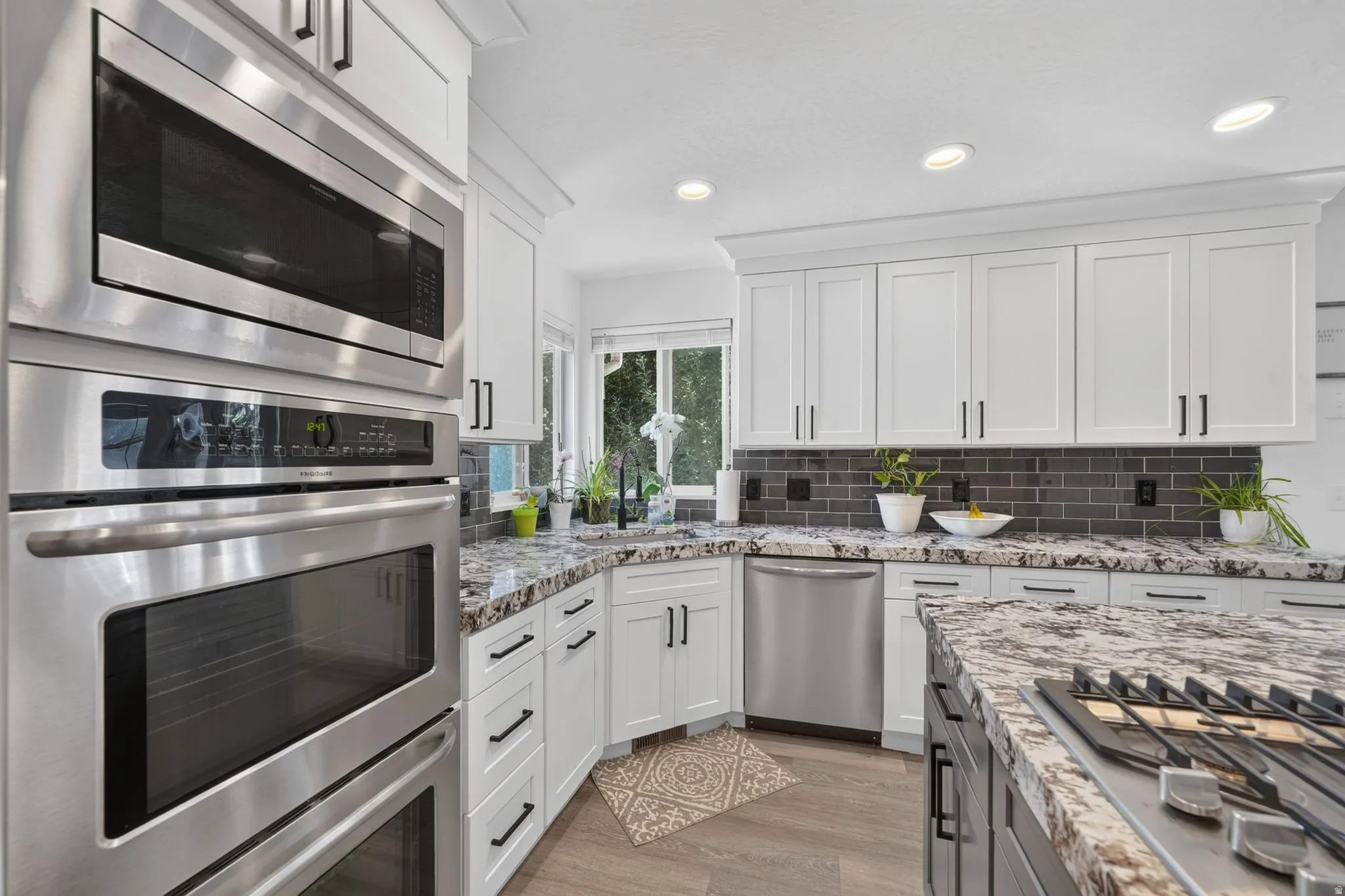White shaker-style cabinetry with matte black hardware, stainless steel built-in appliances, undermount sink, multi-tone stone countertops, and gray subway tile backsplash
