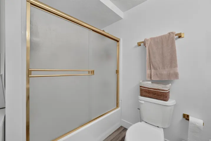 White bathtub with brass-finish sliding glass enclosure, white toilet, brass-finish towel bar, and wood-finish flooring