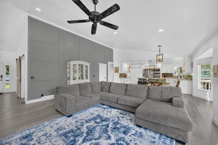 Vaulted ceilings with recessed lighting, wood-finish flooring, accent wall with decorative paneling, black ceiling fan, and a kitchen with white cabinetry