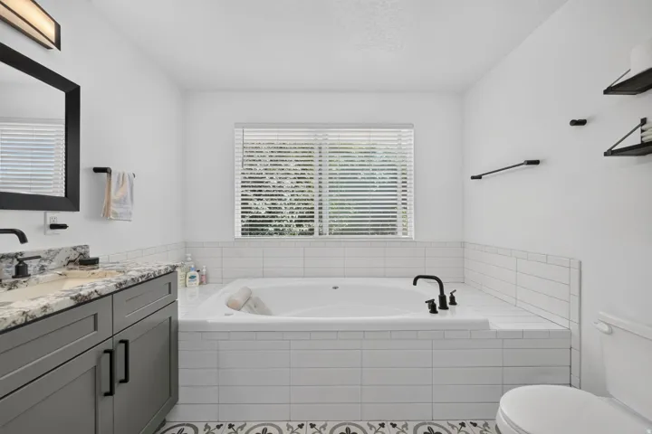 Bathroom featuring a large built-in soaking tub, a single vanity with a stone countertop, a window with blinds, and decorative floor tiling