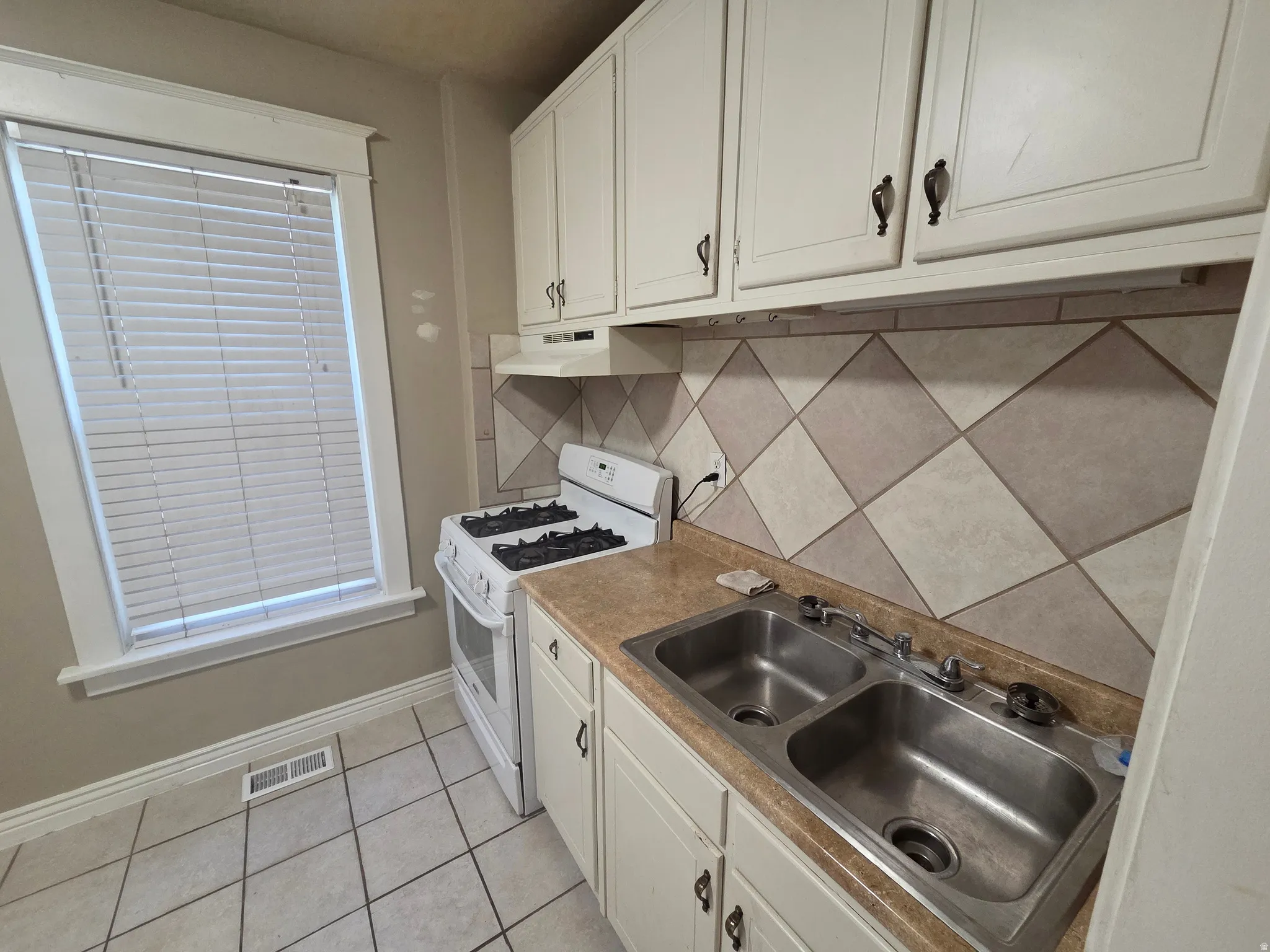 White cabinetry with dark hardware, a dual stainless steel sink, a tiled backsplash, and light-toned flooring