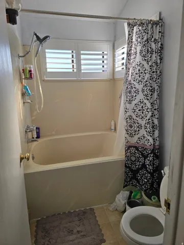 Bathtub with an integrated shower fixture, white louvered window treatments, and neutral-toned wall surfaces