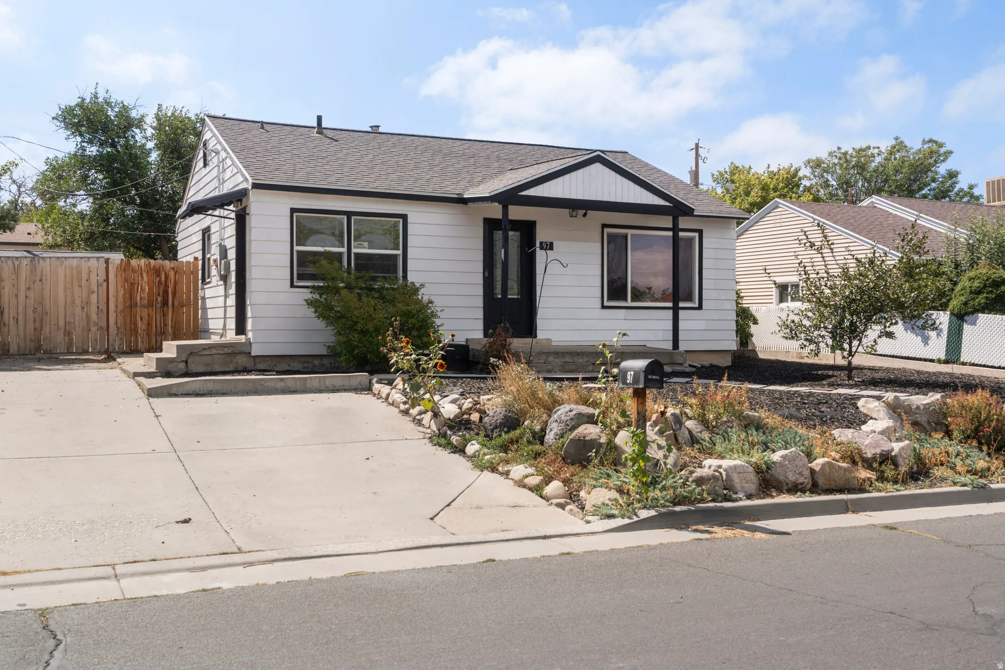 White siding exterior with black trim, concrete driveway, front porch, dark shingle roof, and rock landscape design