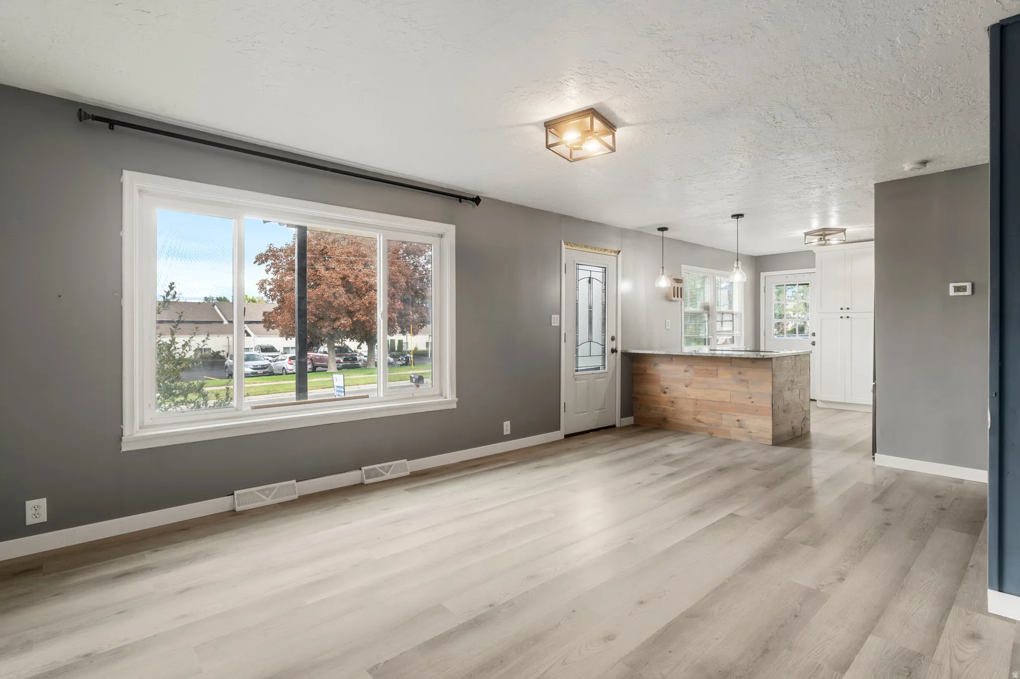 Expansive window with white trim, wood-finish flooring, two modern flush-mount light fixtures, and a peninsula with wood paneling and a light-colored countertop