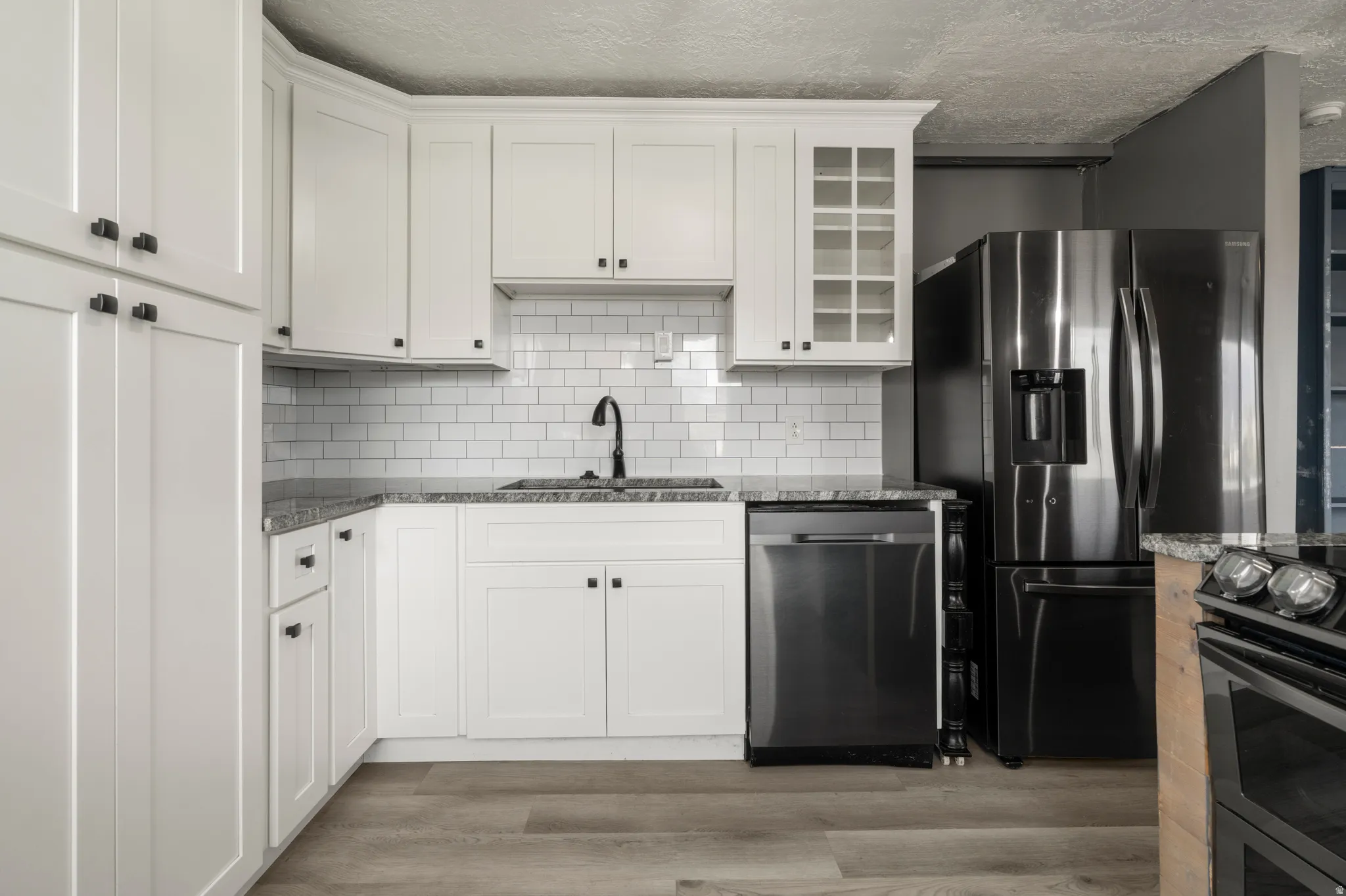 Bright kitchen featuring white shaker cabinetry, stone countertops, a subway tile backsplash, stainless steel appliances, and wood-finish flooring