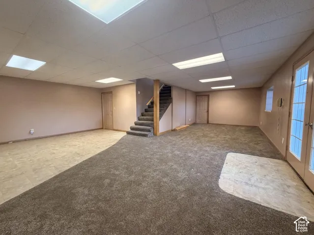 Spacious open basement featuring multiple finished areas with neutral carpet and tile flooring, recessed and panel ceiling lighting, interior staircase with wooden railings, and glass-panel exterior doors providing natural light