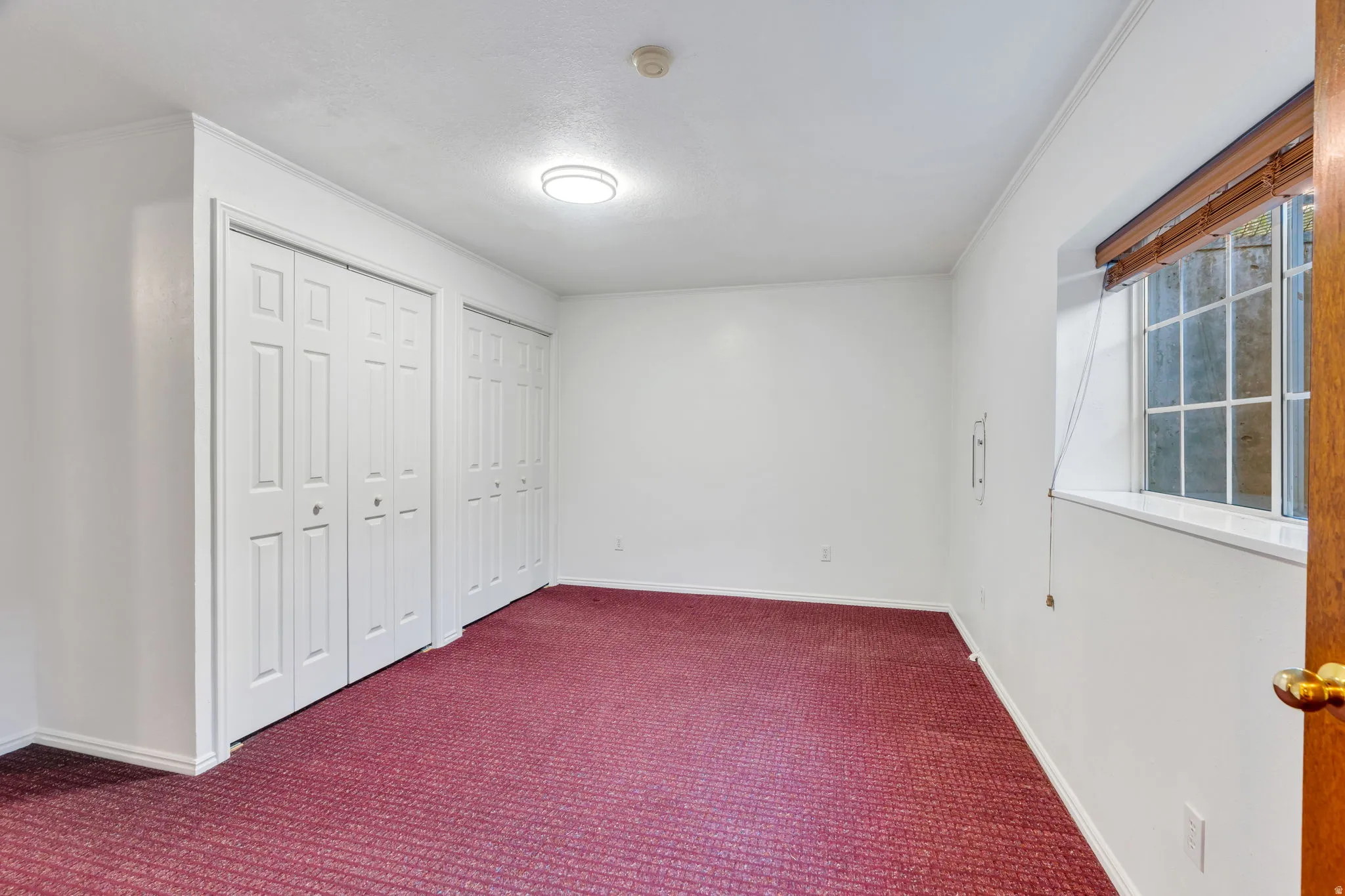 Basement Bedroom: Neutral wall finishes, dual bifold closet doors, patterned carpeting, and a window with wood blinds