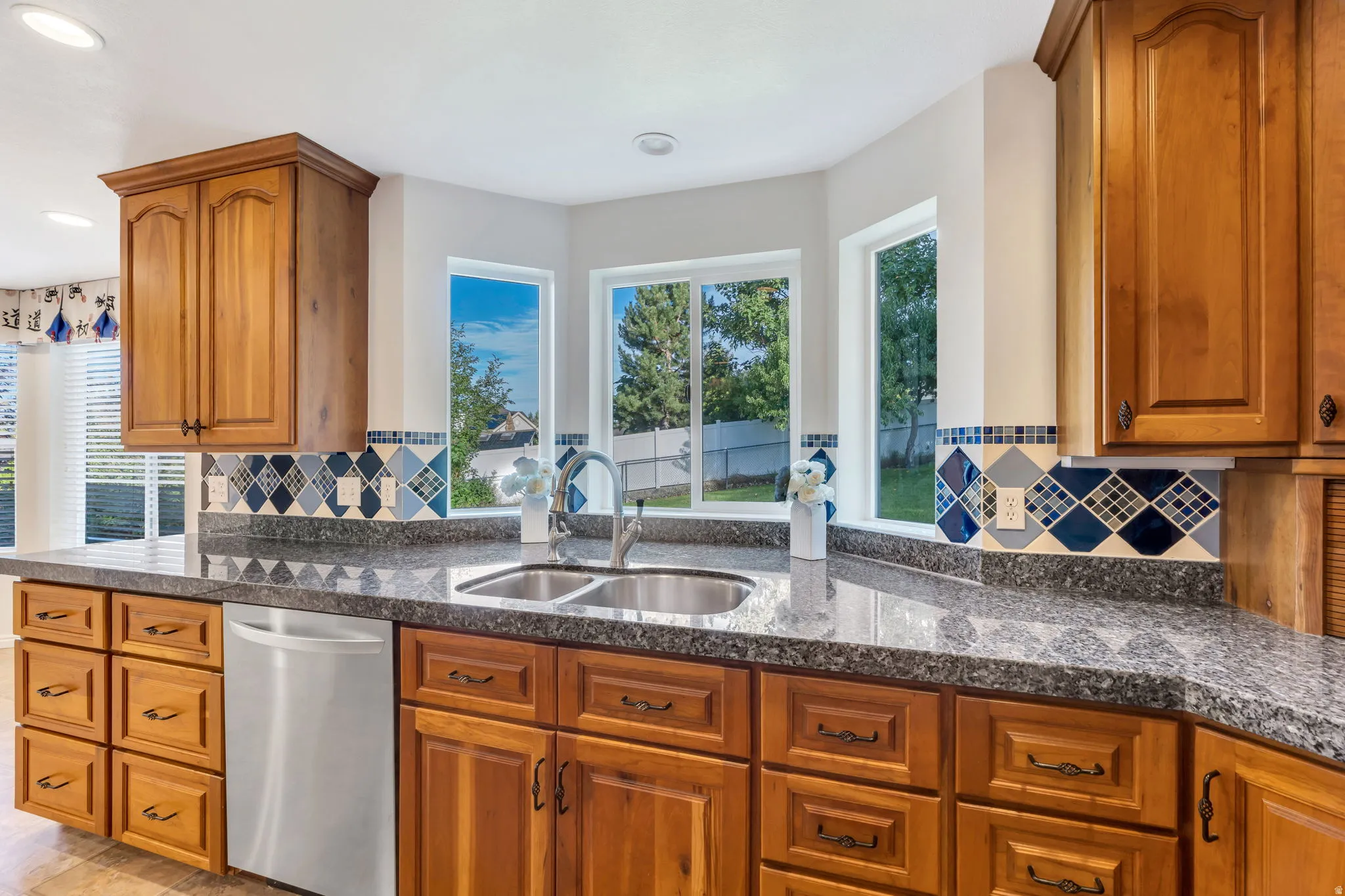 Kitchen featuring natural wood cabinetry, dark speckled countertops, a stainless steel double basin sink with a high-arch faucet, and a patterned tile backsplash