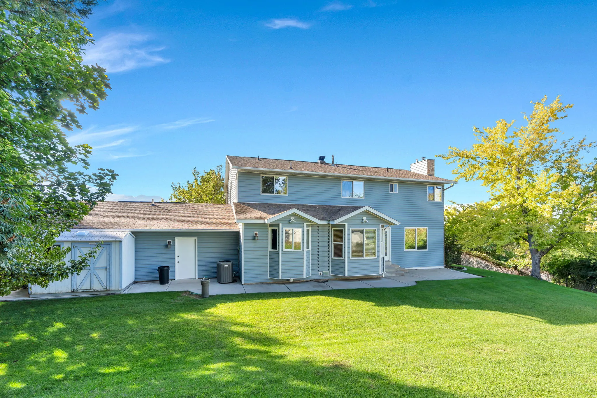 Backyard .35 Acre, Expansive green lawn with a sloped grade, concrete patio area, light blue exterior siding, and a detached shed structure