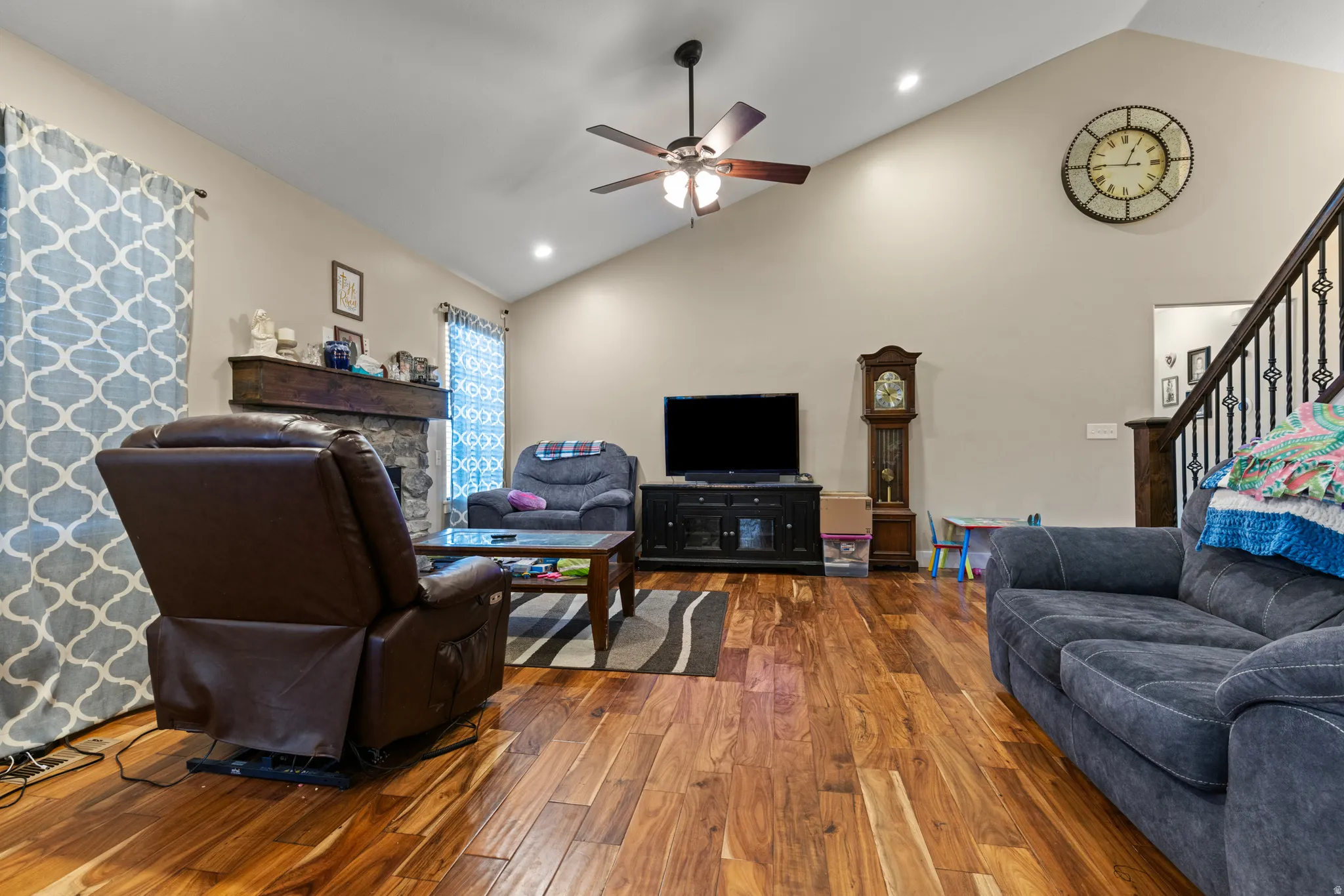 Vaulted ceiling, wood-finish flooring, stone fireplace with wood mantel, ceiling fan with integrated lighting, and wrought iron stair railing