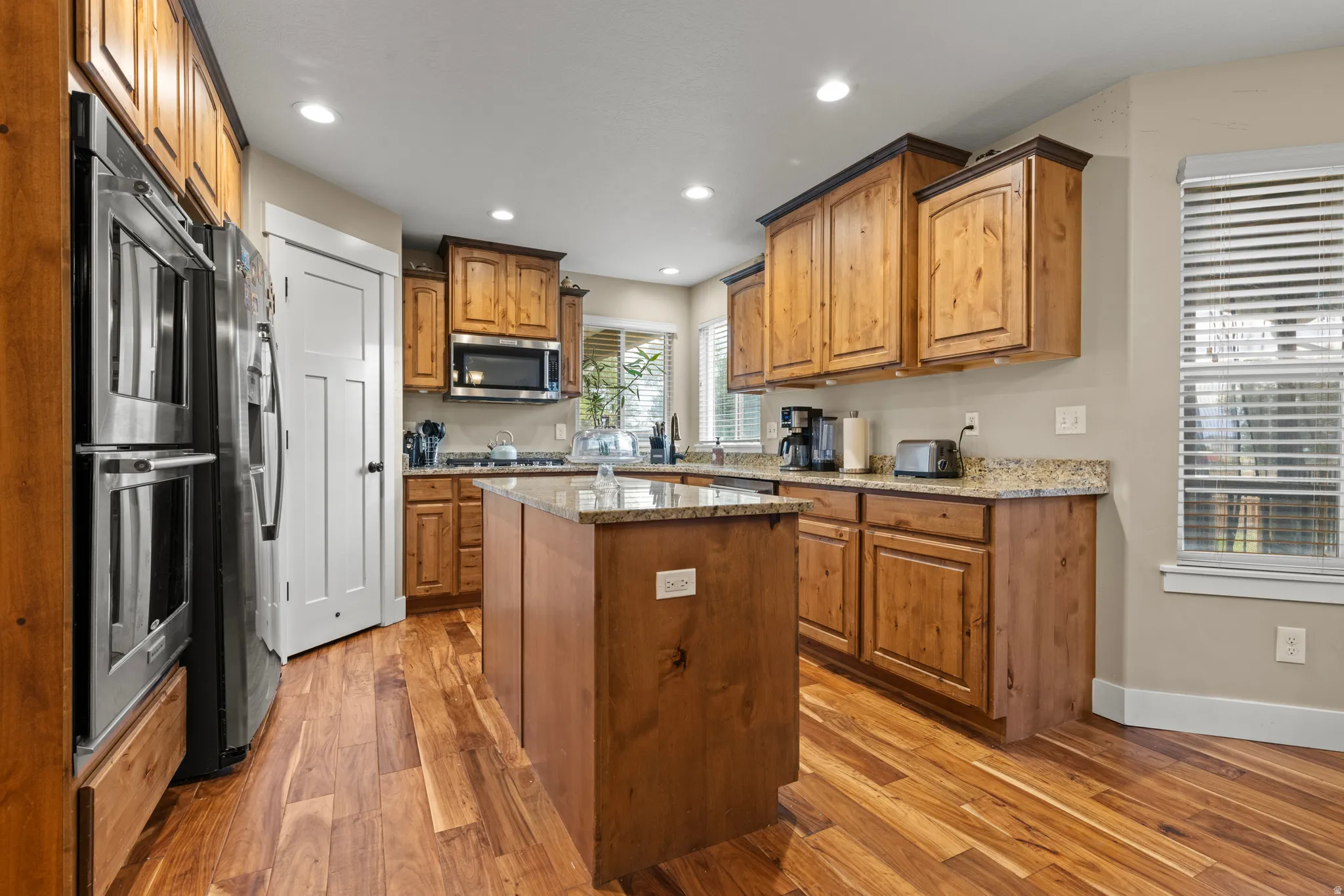 Chef's kitchen featuring warm wood cabinetry, stainless steel appliances including a double wall oven, granite countertops, a substantial center island with seating, recessed lighting, and gleaming hardwood flooring throughout the open layout