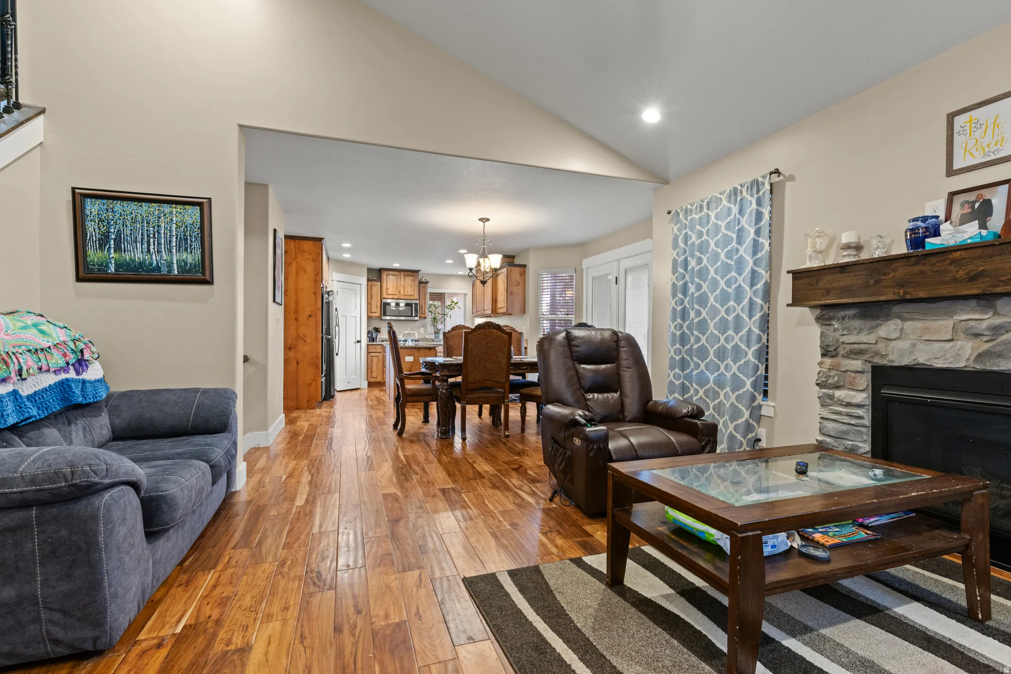 Vaulted ceiling, stone surround fireplace with wood mantle, wood-finish flooring, recessed lighting, and a defined dining area