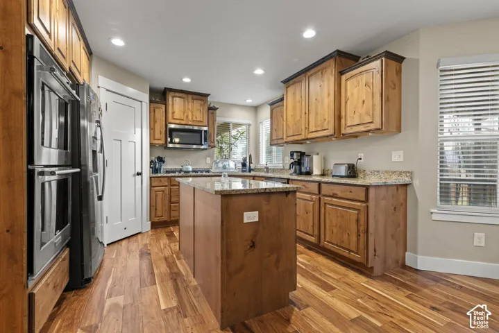 Chef's kitchen featuring warm wood cabinetry, stainless steel appliances including a double wall oven, granite countertops, a substantial center island with seating, recessed lighting, and gleaming hardwood flooring throughout the open layout