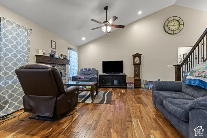 Vaulted ceiling, wood-finish flooring, stone fireplace with wood mantel, ceiling fan with integrated lighting, and wrought iron stair railing