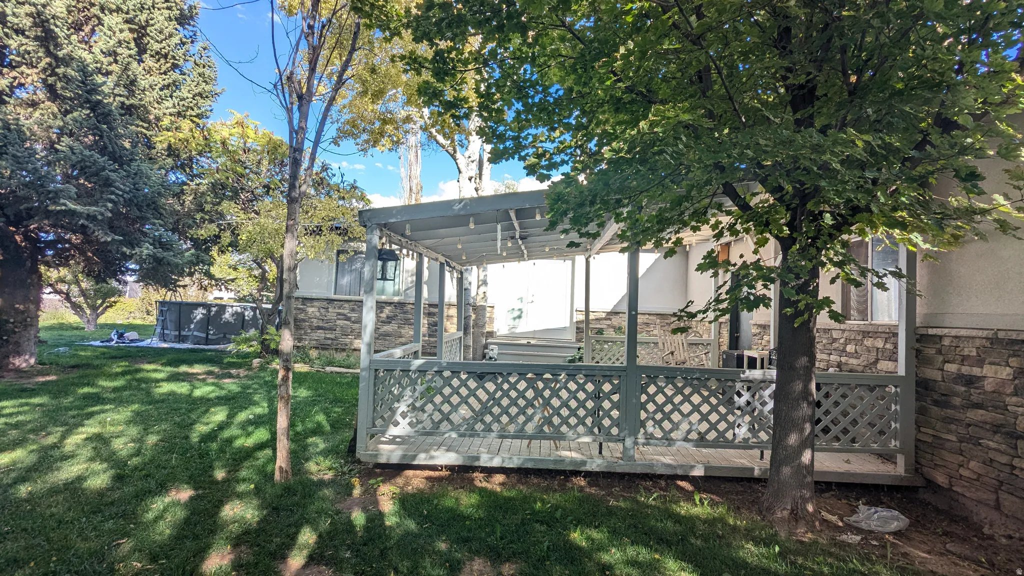 Covered deck with lattice railing, stone veneer foundation, and mature trees