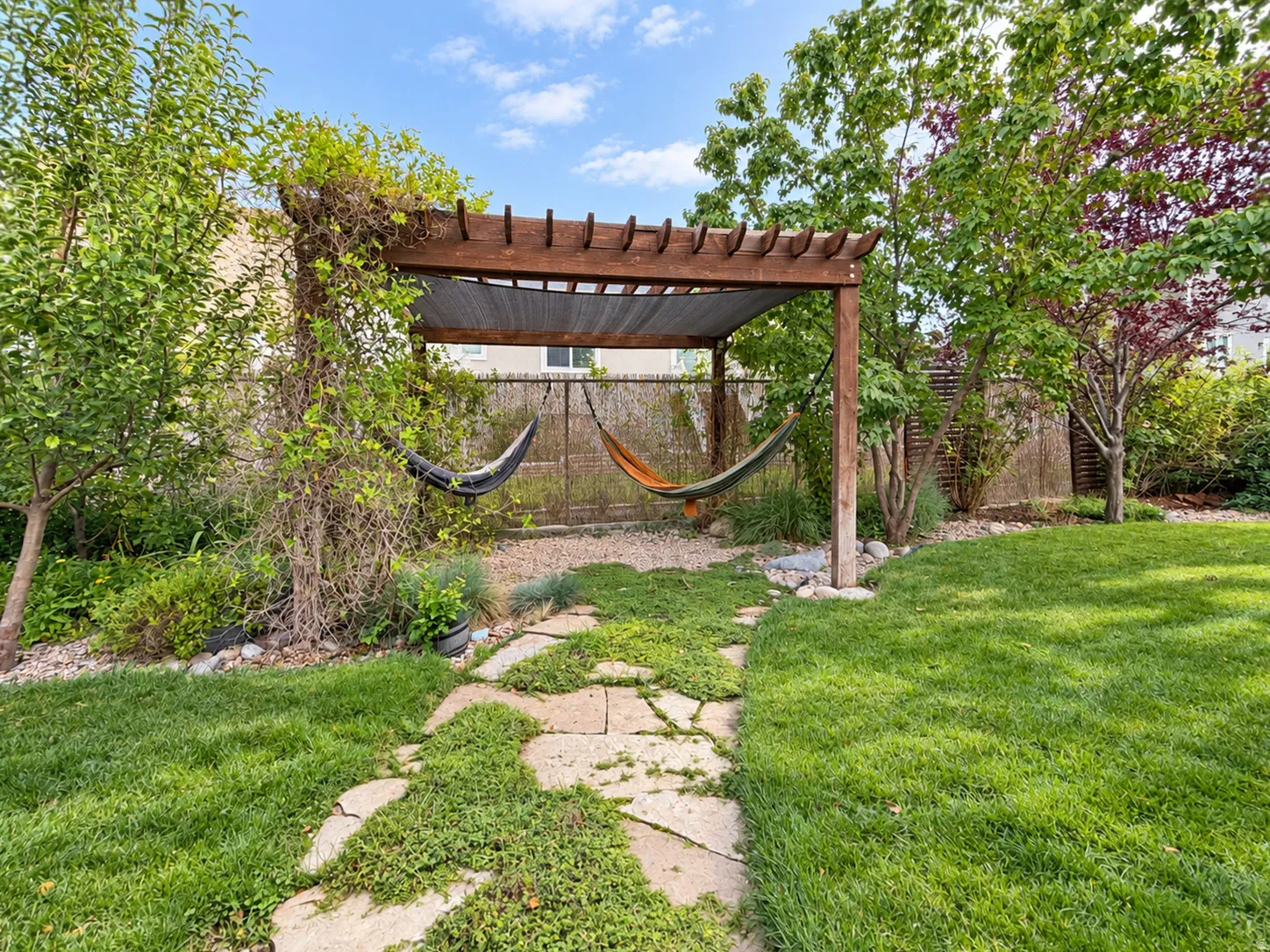 Expansive green lawn with a flagstone pathway, leading to a wood pergola with a shade canopy, surrounded by mature trees and varied plantings