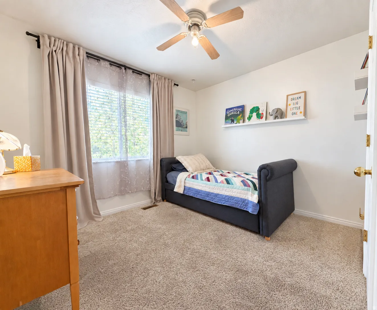 Neutral-toned wall-to-wall carpeting, window with blinds and sheer curtains, ceiling fan with light fixture, and wall-mounted shelving