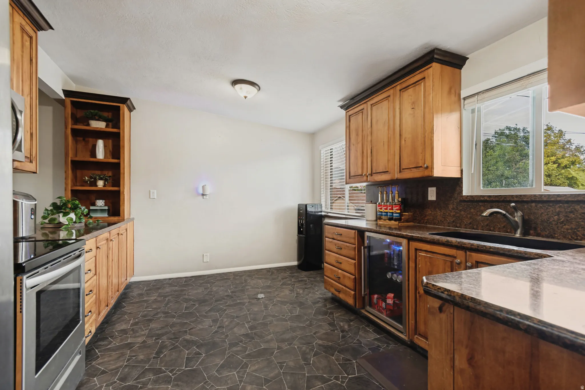 Custom wood cabinetry with dark crown molding, stone-finish flooring, stainless steel range, built-in beverage cooler, and dark stone countertops