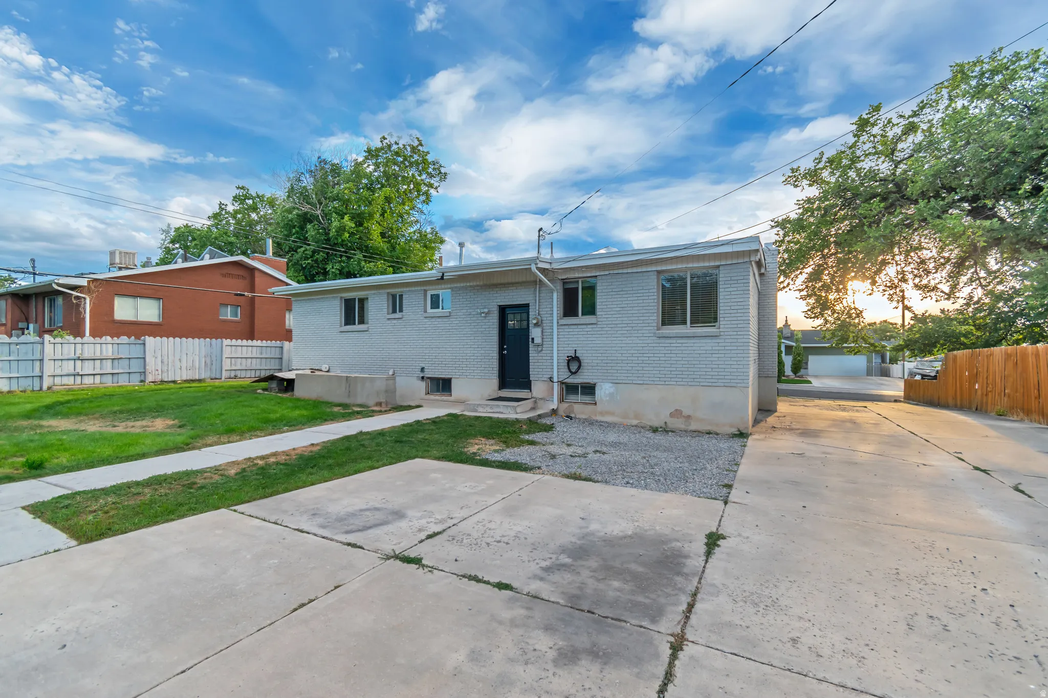 Grey brick exterior with multiple windows, concrete driveway, concrete walk, gravel area, and a dark entry door