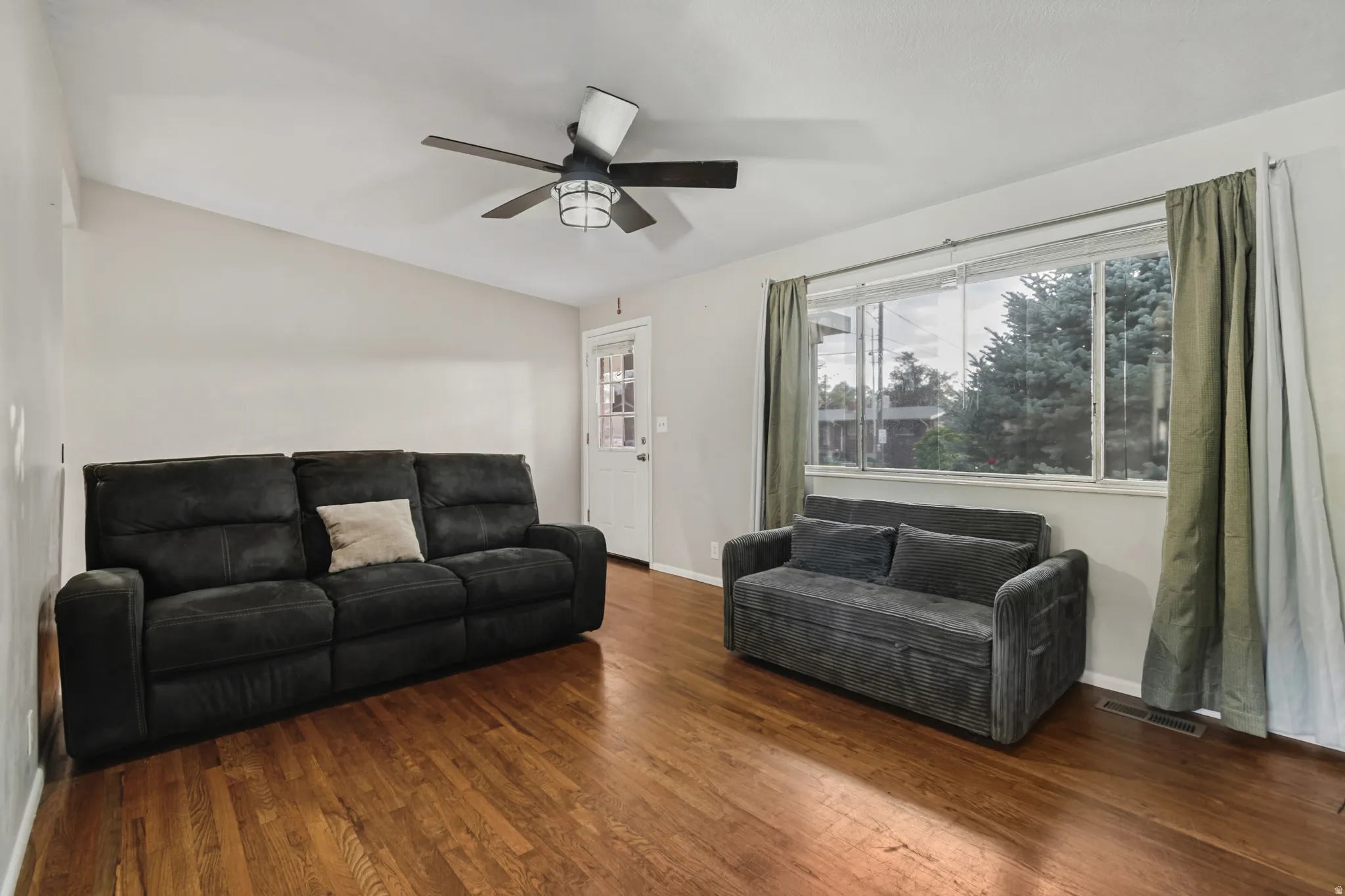 Wood-finish flooring, white walls, large window, ceiling fan with integrated lighting, and a white paneled door