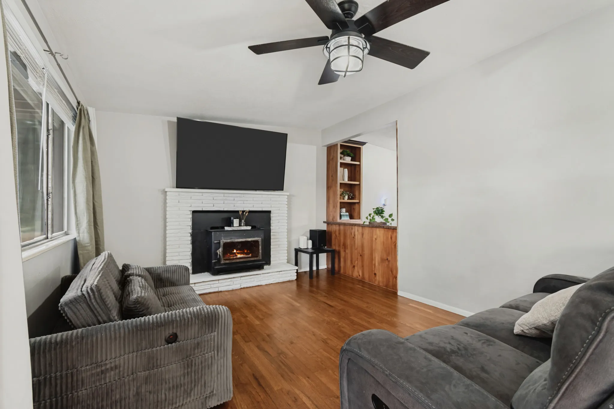 Hardwood flooring, brick fireplace with wood-burning stove insert, built-in shelving, wood paneling, and a ceiling fan with integrated lighting