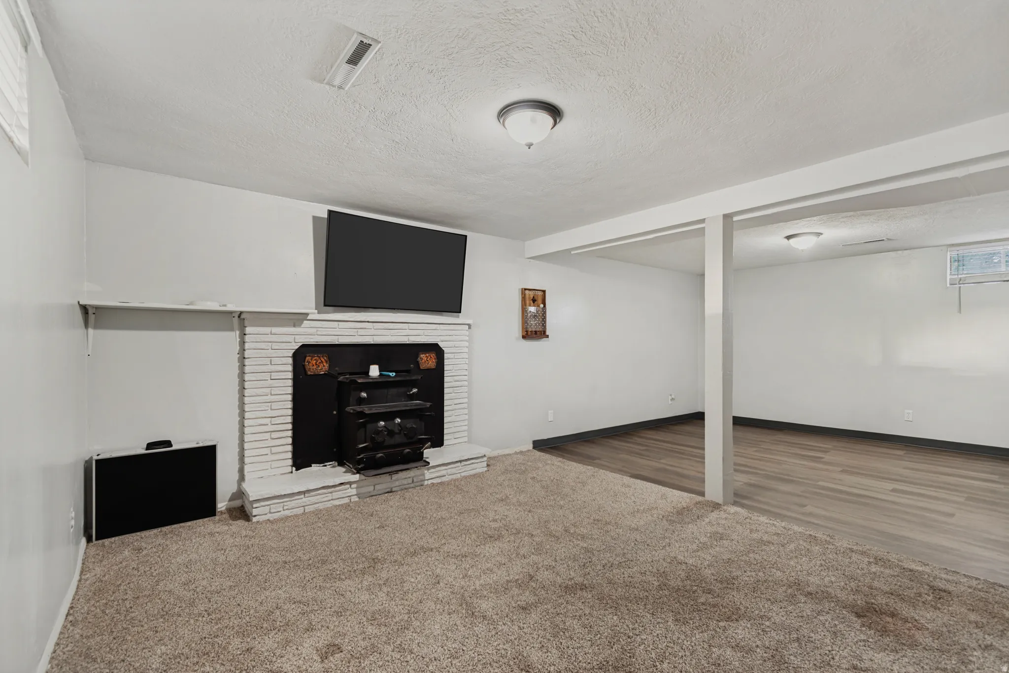 Carpeted room featuring a painted brick fireplace with a hearth, a wall-mounted display, and an adjacent area with wood-finish flooring