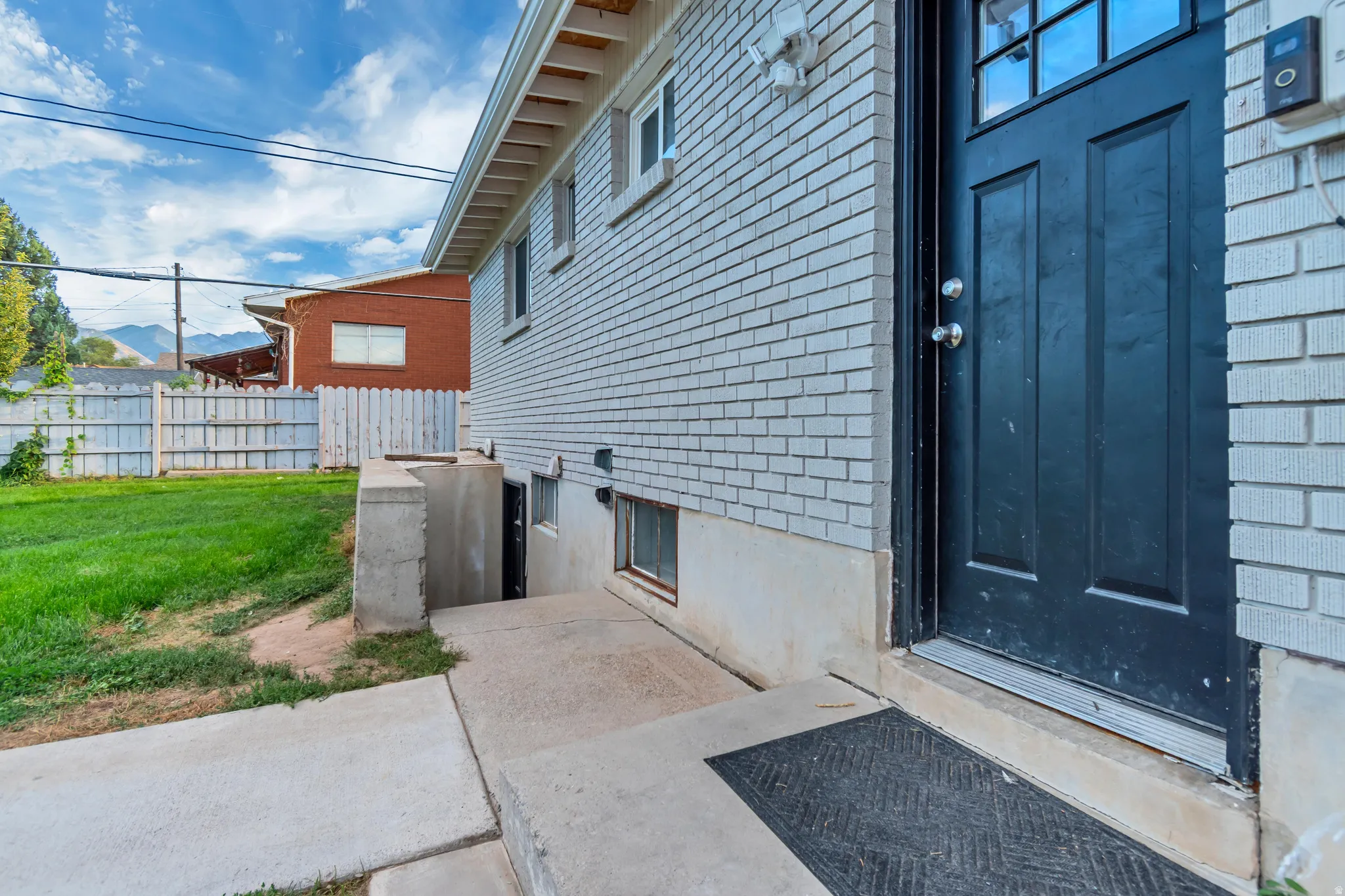Brick exterior with a dark-paneled door, concrete walkway, and a concrete-lined basement access