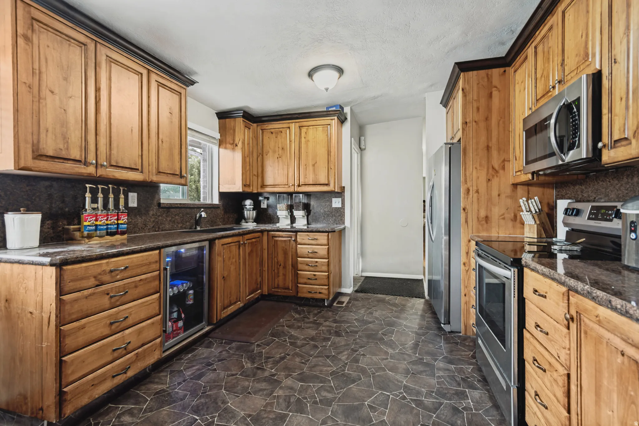 Wood-finish cabinetry with contrasting trim, stone-finish countertops, stainless steel appliances, and a built-in beverage refrigerator