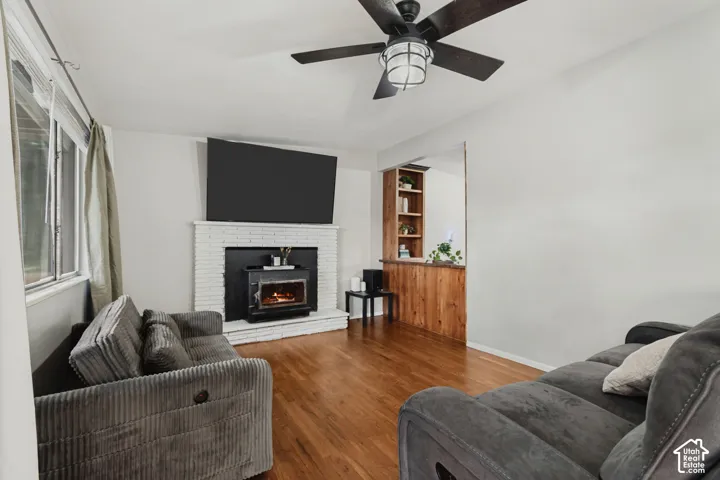 Hardwood flooring, brick fireplace with wood-burning stove insert, built-in shelving, wood paneling, and a ceiling fan with integrated lighting