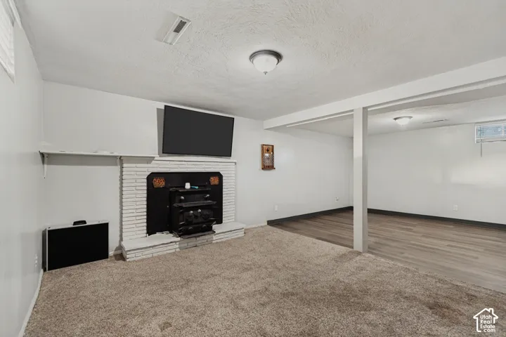Carpeted room featuring a painted brick fireplace with a hearth, a wall-mounted display, and an adjacent area with wood-finish flooring