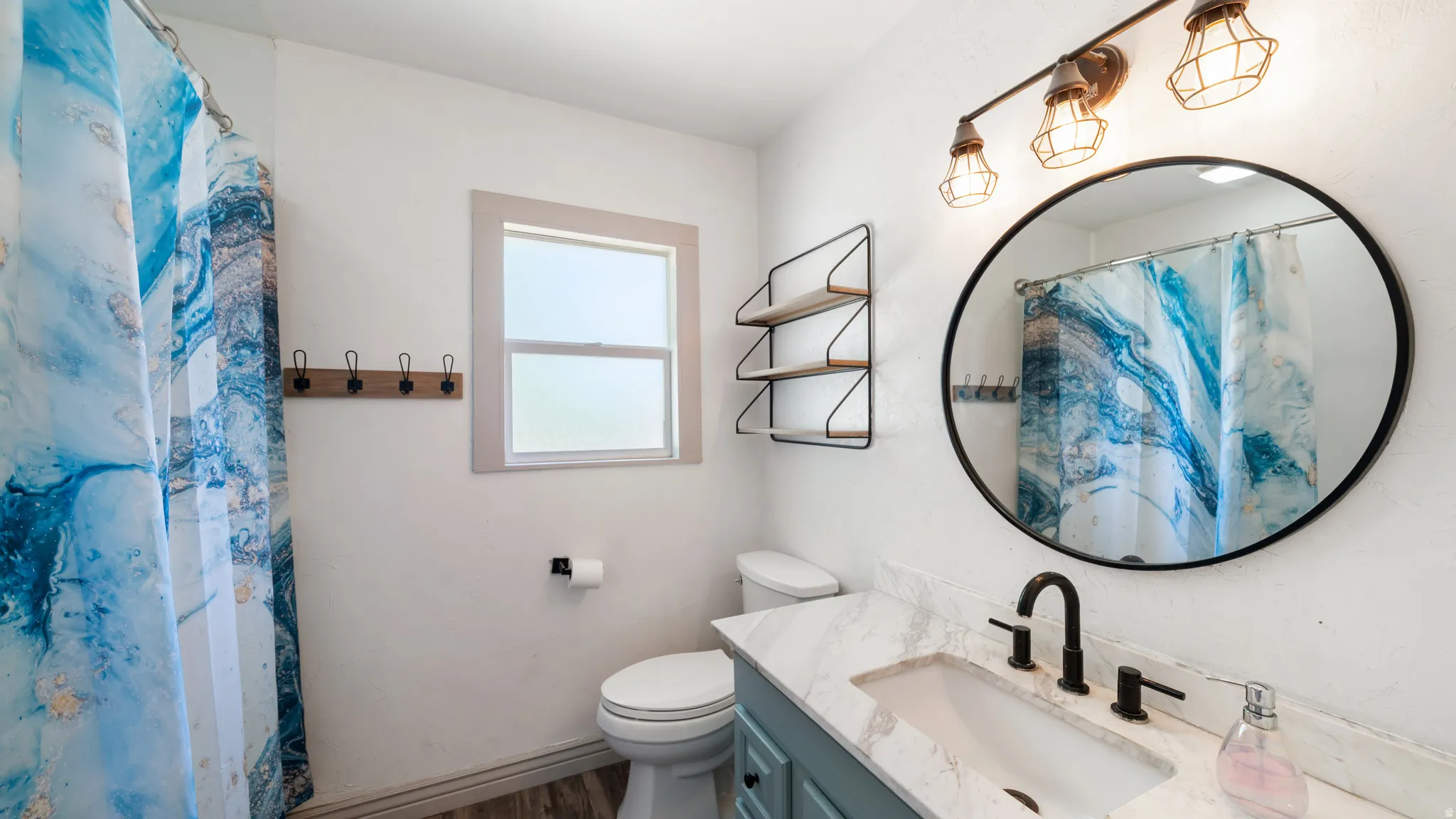 Bathroom featuring a single window, a light blue vanity, a white undermount sink, and a circular wall mirror with an industrial-style vanity light