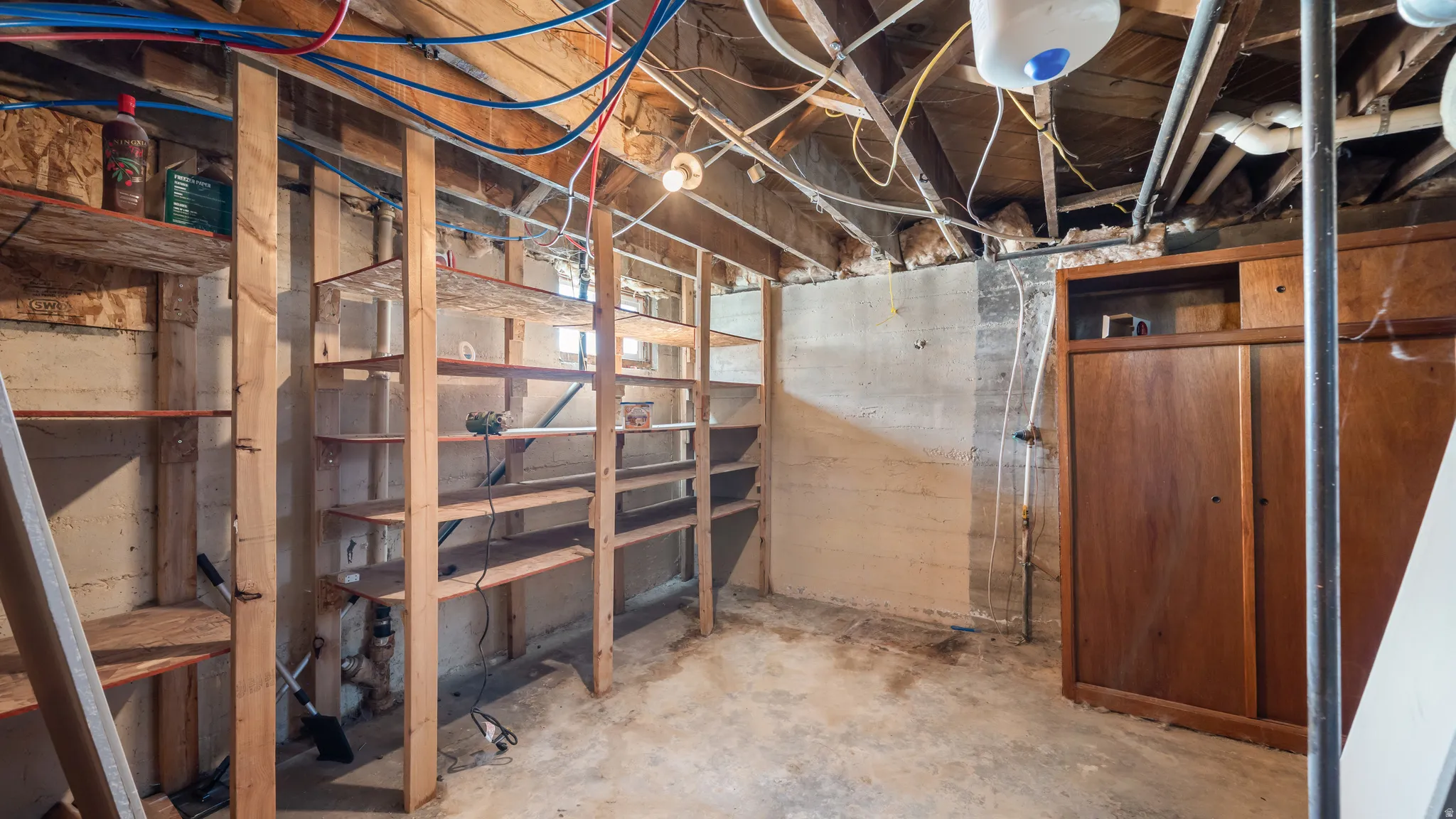Unfinished basement space featuring exposed wood joists, concrete walls, built-in wooden shelving, and a built-in wooden cabinet