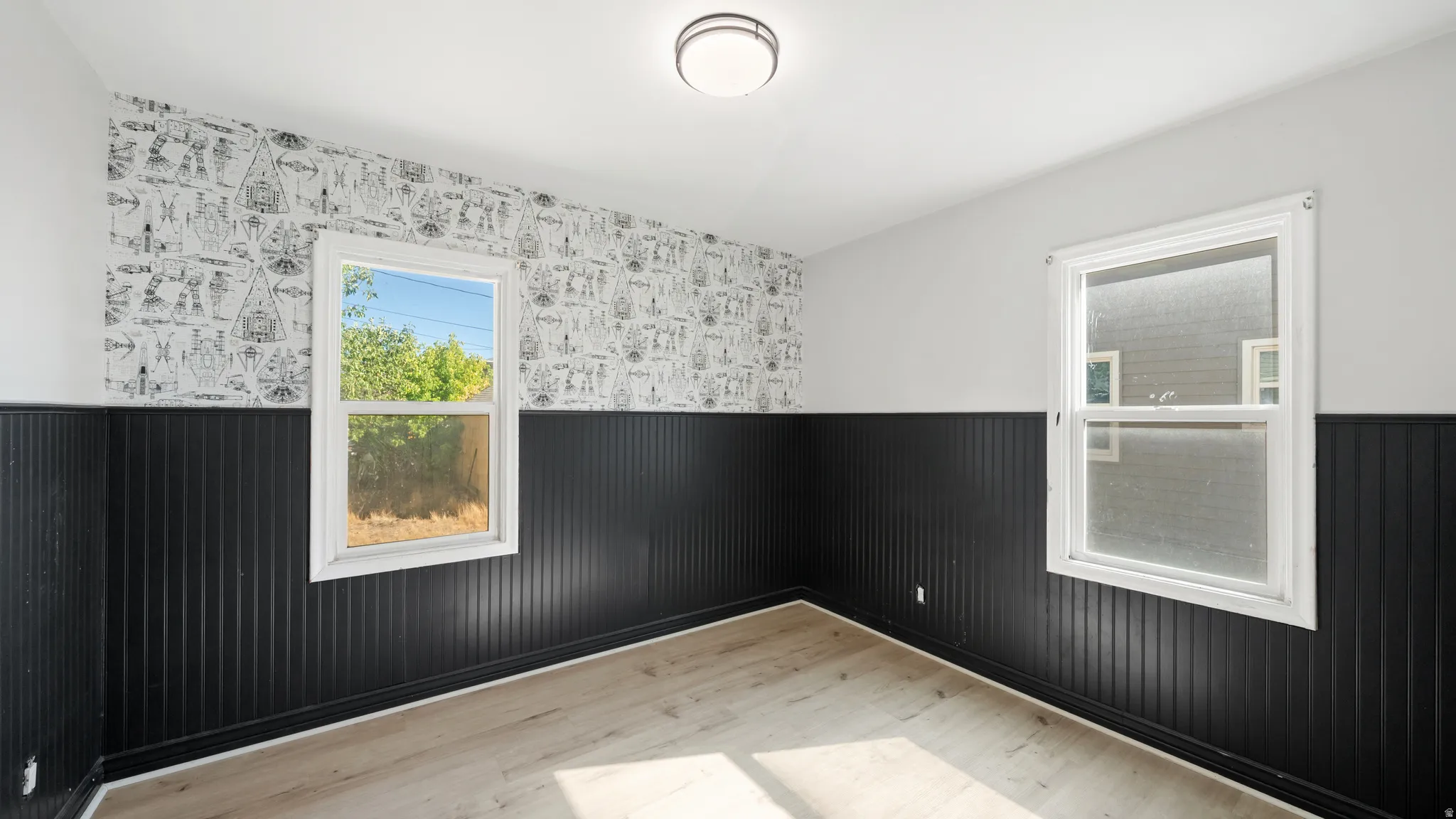 Light wood-finish flooring, two large windows, black vertical paneling, patterned wall covering, and a flush mount ceiling light