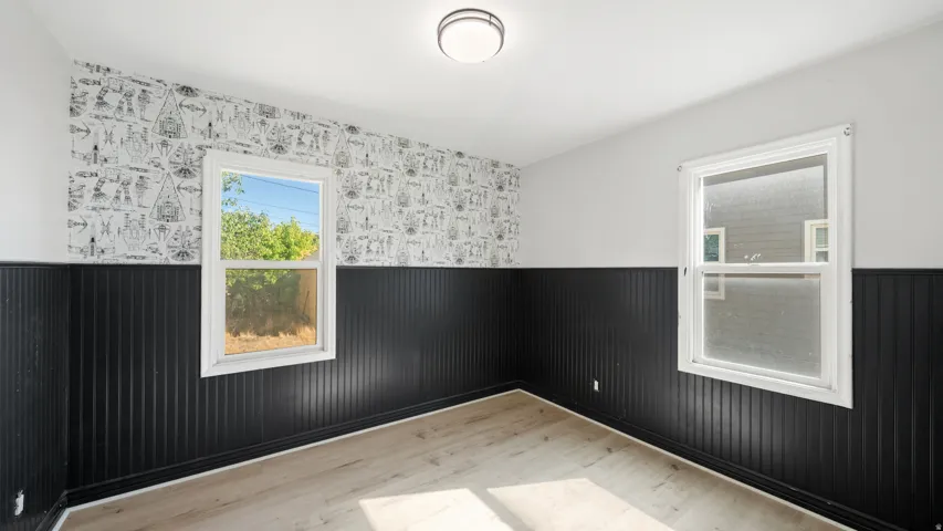 Light wood-finish flooring, two large windows, black vertical paneling, patterned wall covering, and a flush mount ceiling light