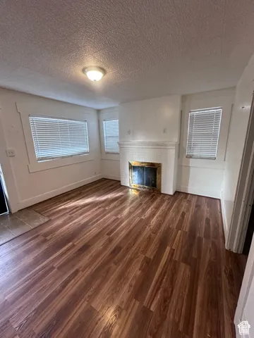 Wood-finish flooring, white tiled fireplace with gold-toned surround, multiple windows with blinds, and a flush mount ceiling light