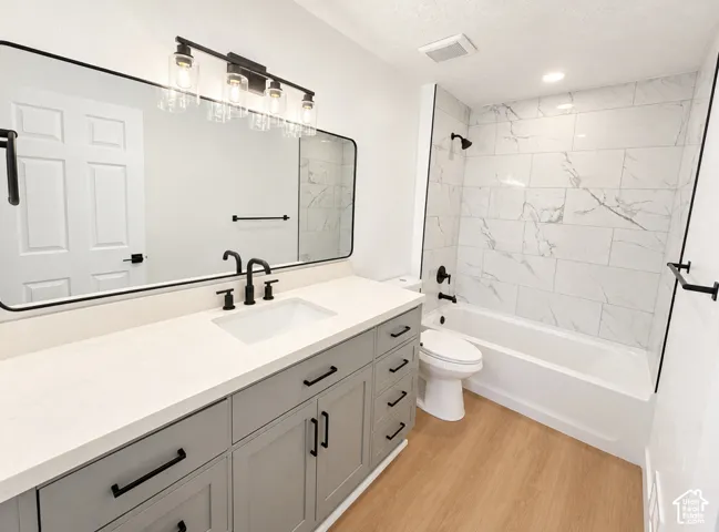 Light wood-finish flooring, a vanity with a light countertop and dark hardware, a rectangular sink with dark fixtures, a large mirror, and a tiled shower/tub combination with dark fixtures