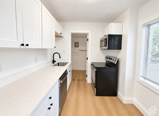 White shaker-style cabinetry, light-toned countertops, stainless steel sink with a matte black faucet, wood-finish flooring, and a window with horizontal blinds