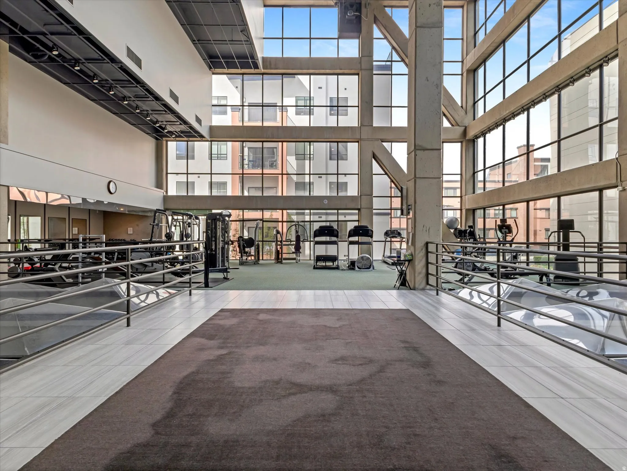 Expansive fitness area with a high ceiling, large glass panels, exposed structural beams, and a combination of carpet and tiled flooring