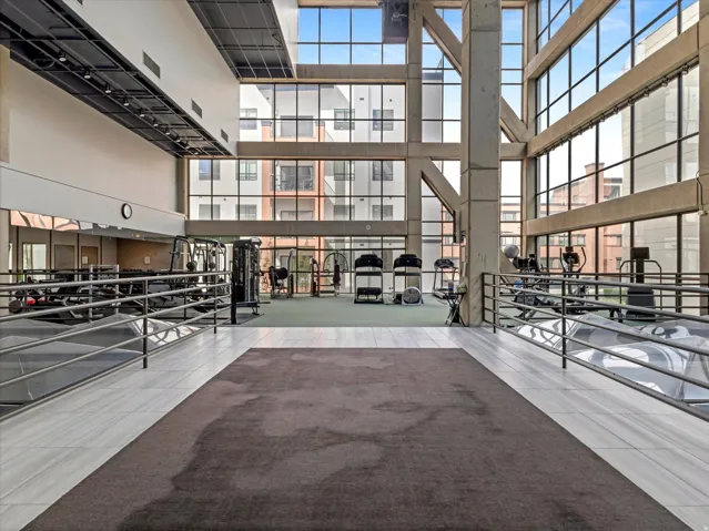 Expansive fitness area with a high ceiling, large glass panels, exposed structural beams, and a combination of carpet and tiled flooring