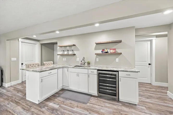 L-shaped wet bar featuring white cabinetry, light-toned countertops, a stainless steel beverage refrigerator, and wood-finish open shelving