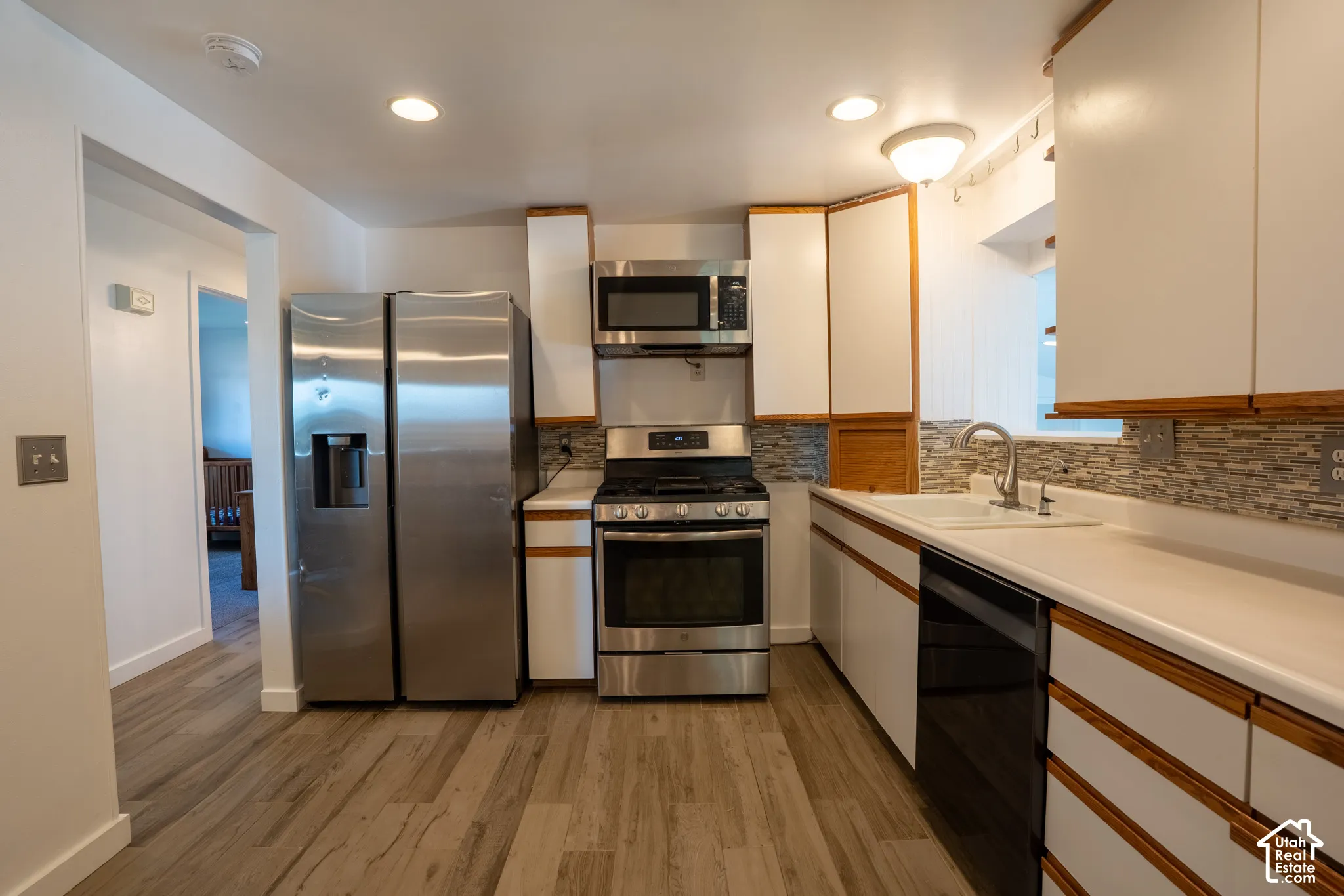 Kitchen featuring wood-finish flooring, white cabinetry with wood trim, stainless steel appliances, a mosaic tile backsplash, and light-toned countertops