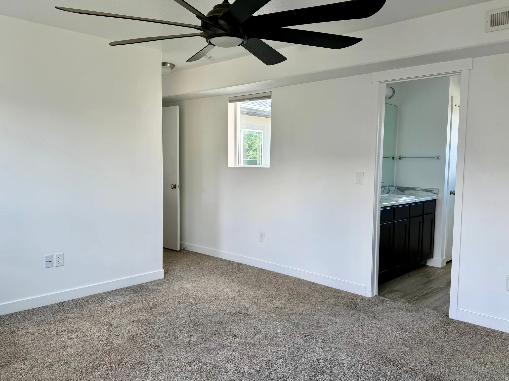 Ceiling fan with five dark blades, window with white trim, light-colored carpet flooring, white walls, and a bathroom entry featuring a dark vanity with a light-colored countertop