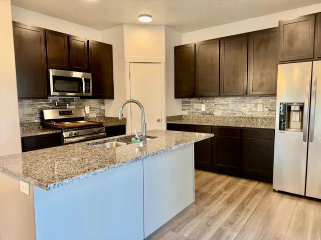 Kitchen featuring an island with a stainless steel sink, dark wood-finish cabinetry, light-toned wood-finish flooring, and a patterned tile backsplash