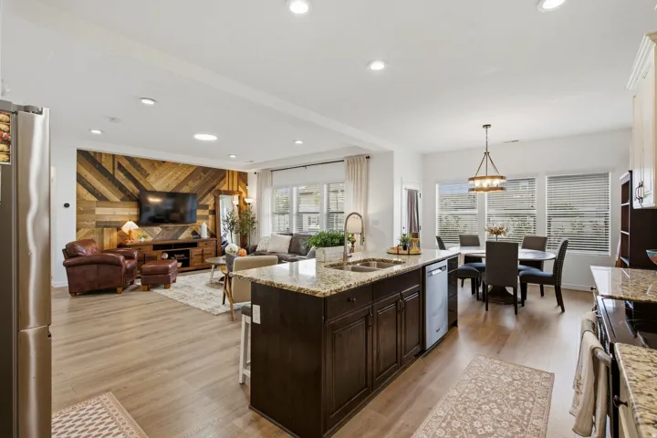 Open-concept living space with wood-finish flooring, a kitchen island with stone countertops, stainless steel appliances, a dining area with a chandelier, and a wood-paneled accent wall