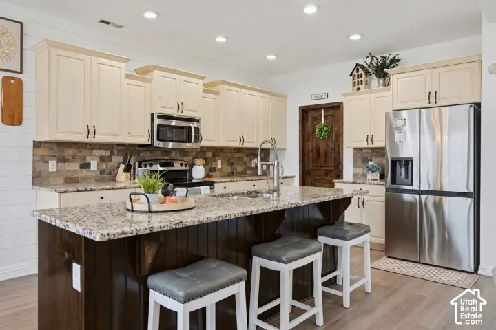 Kitchen featuring a large central island with seating, light-toned upper cabinetry, stainless steel appliances, stacked stone backsplash, and wood-finish flooring