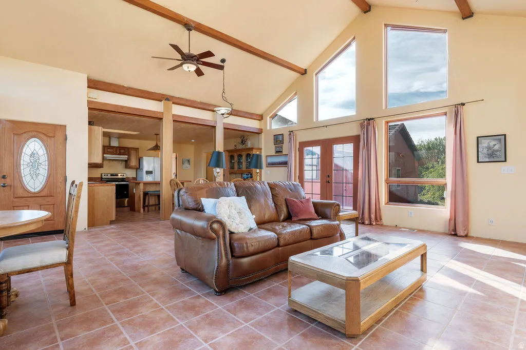 Open-concept living area featuring vaulted ceilings with exposed wooden beams, ceiling fan, and abundant natural light from multiple large windows and glass doors